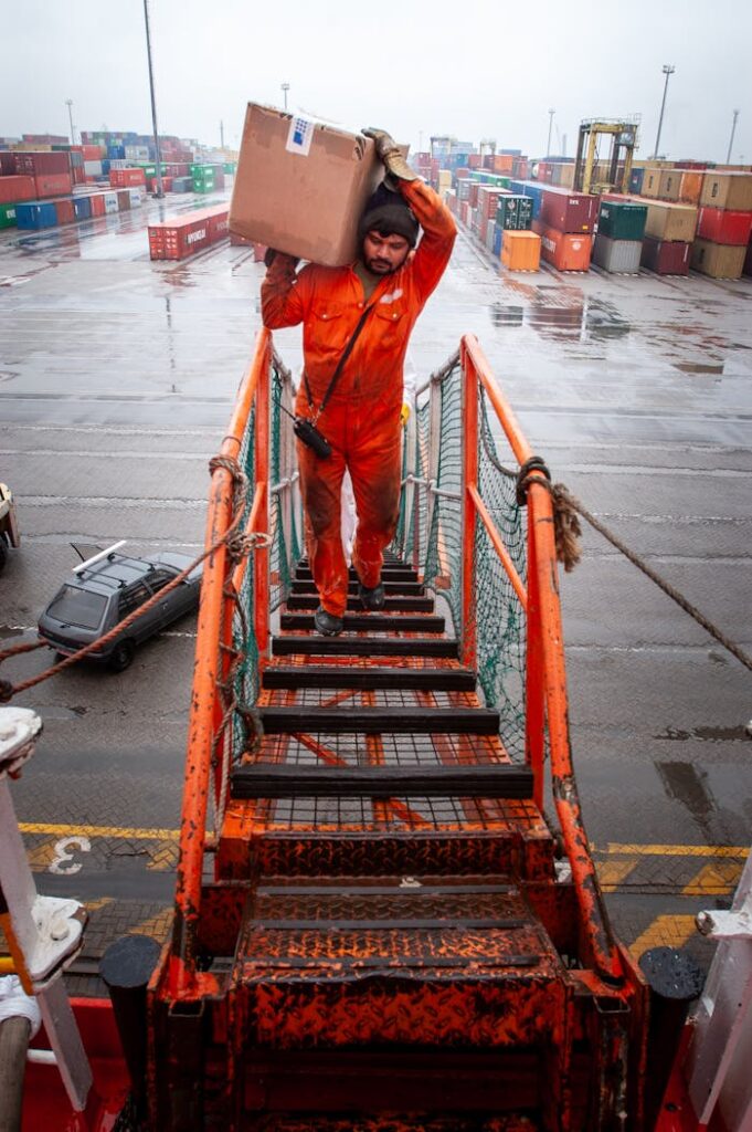 A maritime worker in orange coveralls carries a box up a ship's gangway in rainy weather at a port.
